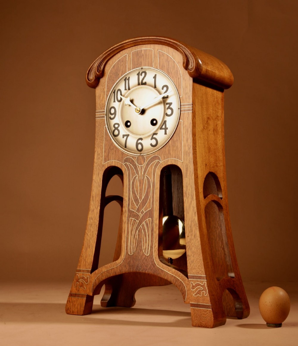 An Art Nouveau oak mantel clock featuring stylised inlaid line decoration, an arched top, and an open lower structure exposing the pendulum.