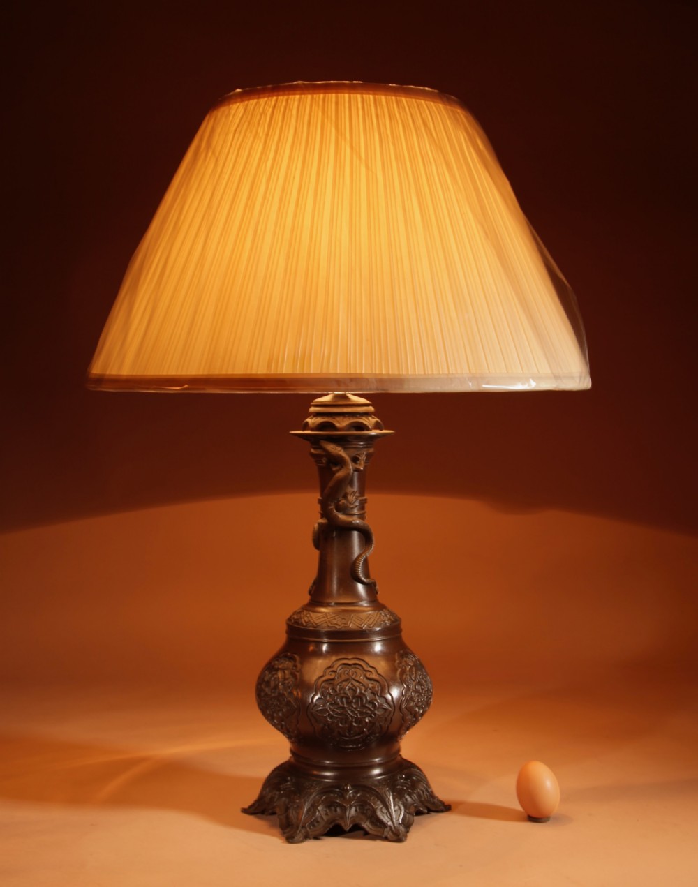 A dark patinated metal table lamp with relief dragon decoration, fitted with a pleated flared lampshade.