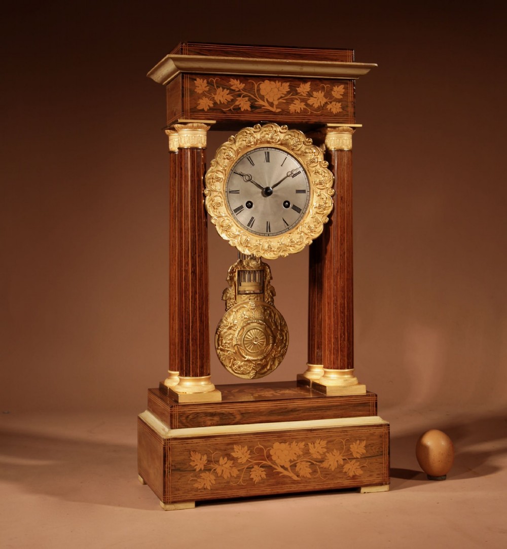 An antique rosewood portico clock with four fluted columns, gilt ormolu capitals, floral lemonwood marquetry inlays, and an exposed pendulum.