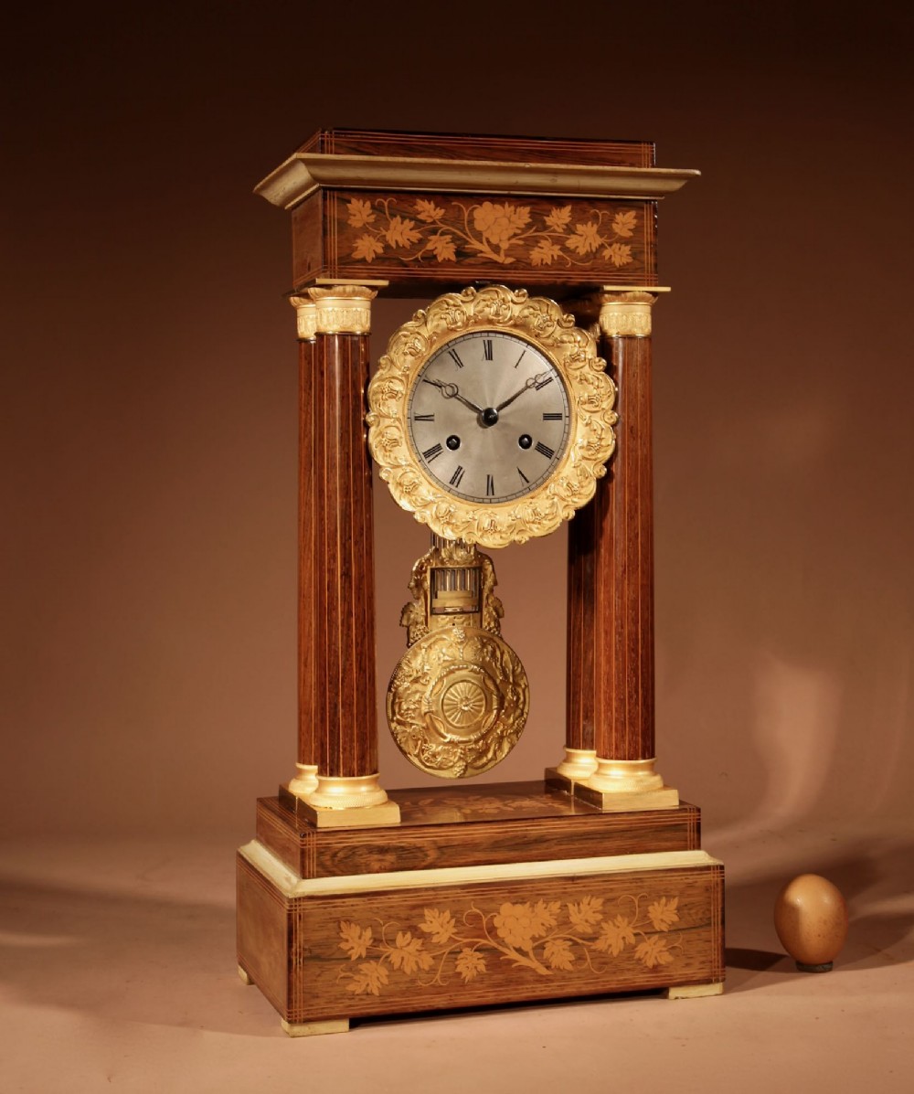 An antique rosewood portico clock with four fluted columns, gilt ormolu capitals, floral lemonwood marquetry inlays, and an exposed pendulum. View II