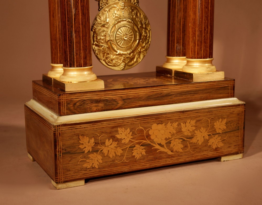 An antique rosewood portico clock with four fluted columns, gilt ormolu capitals, floral lemonwood marquetry inlays, and an exposed pendulum. View III