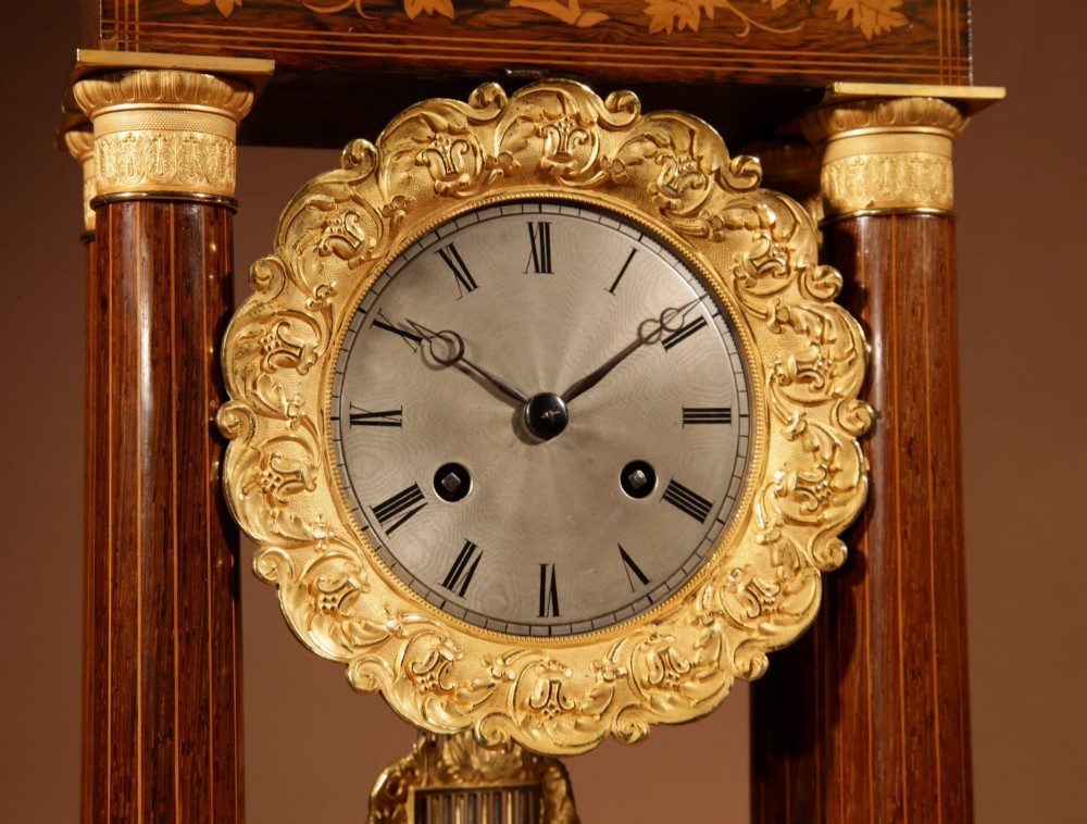An antique rosewood portico clock with four fluted columns, gilt ormolu capitals, floral lemonwood marquetry inlays, and an exposed pendulum. View IV