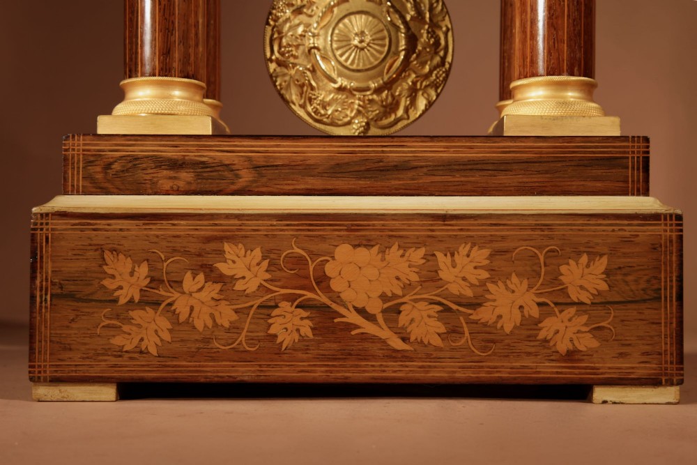 An antique rosewood portico clock with four fluted columns, gilt ormolu capitals, floral lemonwood marquetry inlays, and an exposed pendulum. View V