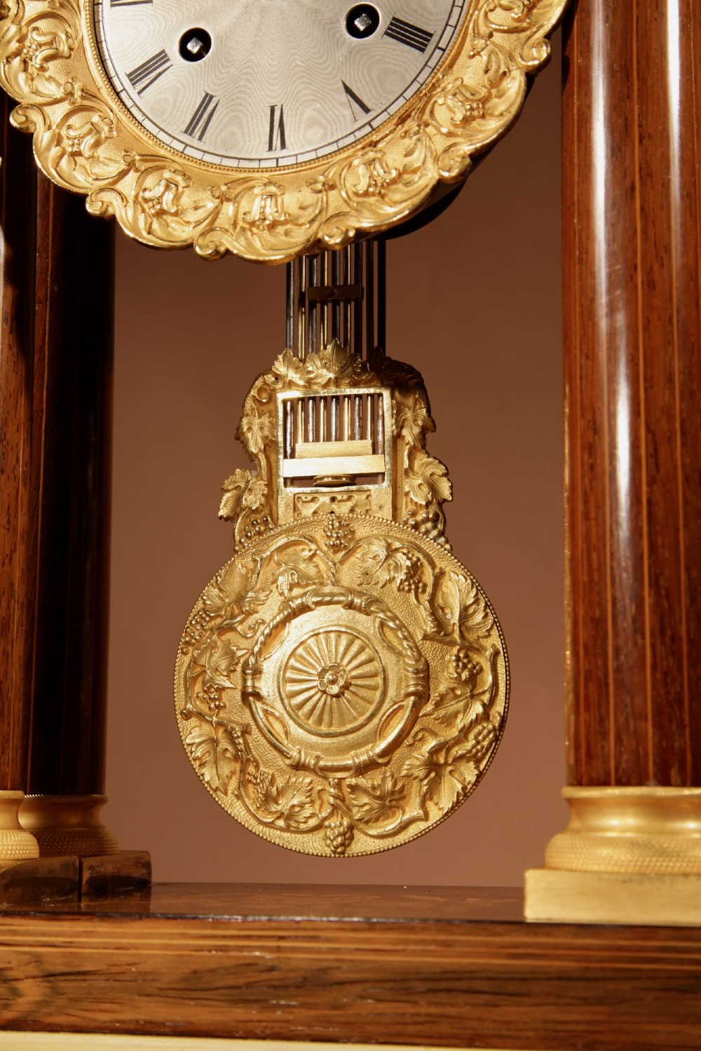 An antique rosewood portico clock with four fluted columns, gilt ormolu capitals, floral lemonwood marquetry inlays, and an exposed pendulum. View VI