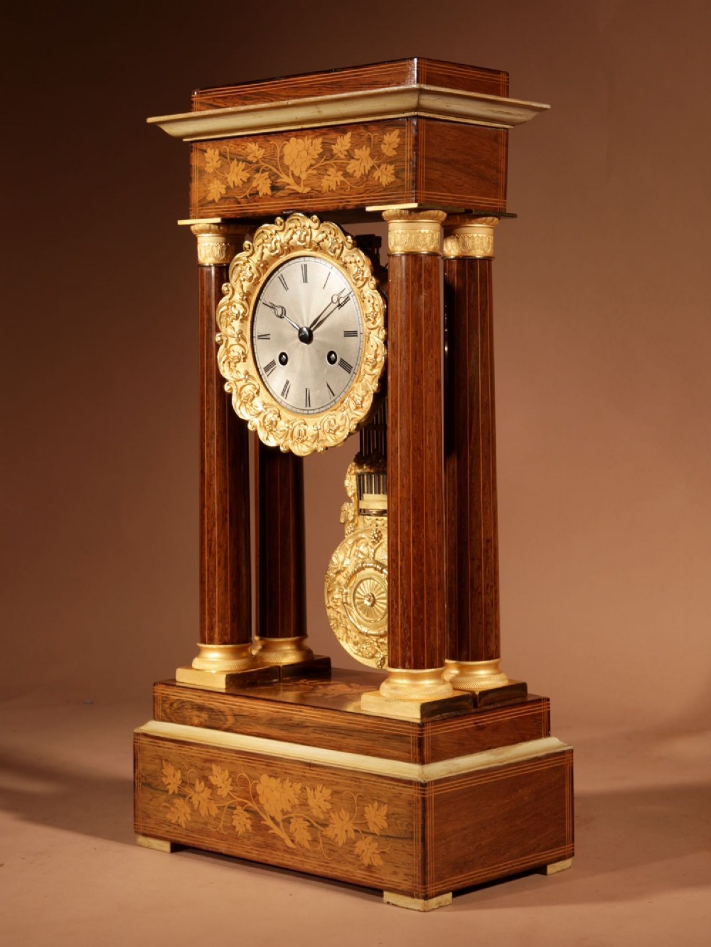 An antique rosewood portico clock with four fluted columns, gilt ormolu capitals, floral lemonwood marquetry inlays, and an exposed pendulum. View VII