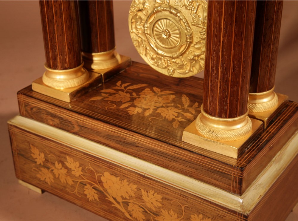 An antique rosewood portico clock with four fluted columns, gilt ormolu capitals, floral lemonwood marquetry inlays, and an exposed pendulum. View VIII