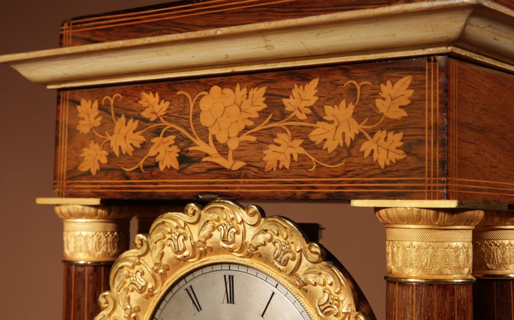 An antique rosewood portico clock with four fluted columns, gilt ormolu capitals, floral lemonwood marquetry inlays, and an exposed pendulum. View IX