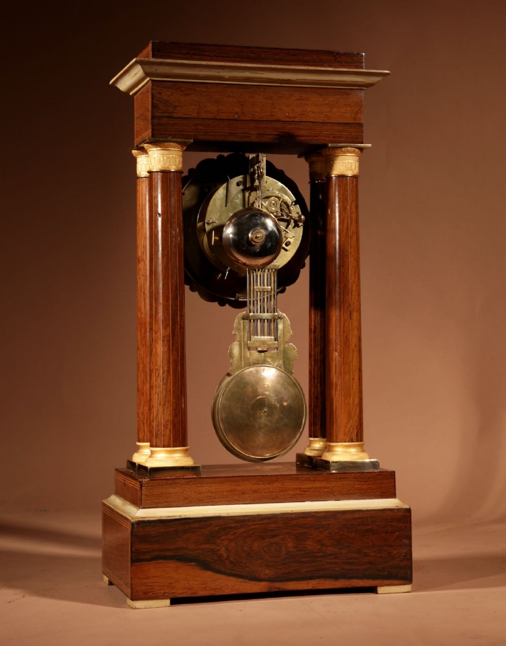 An antique rosewood portico clock with four fluted columns, gilt ormolu capitals, floral lemonwood marquetry inlays, and an exposed pendulum. View X