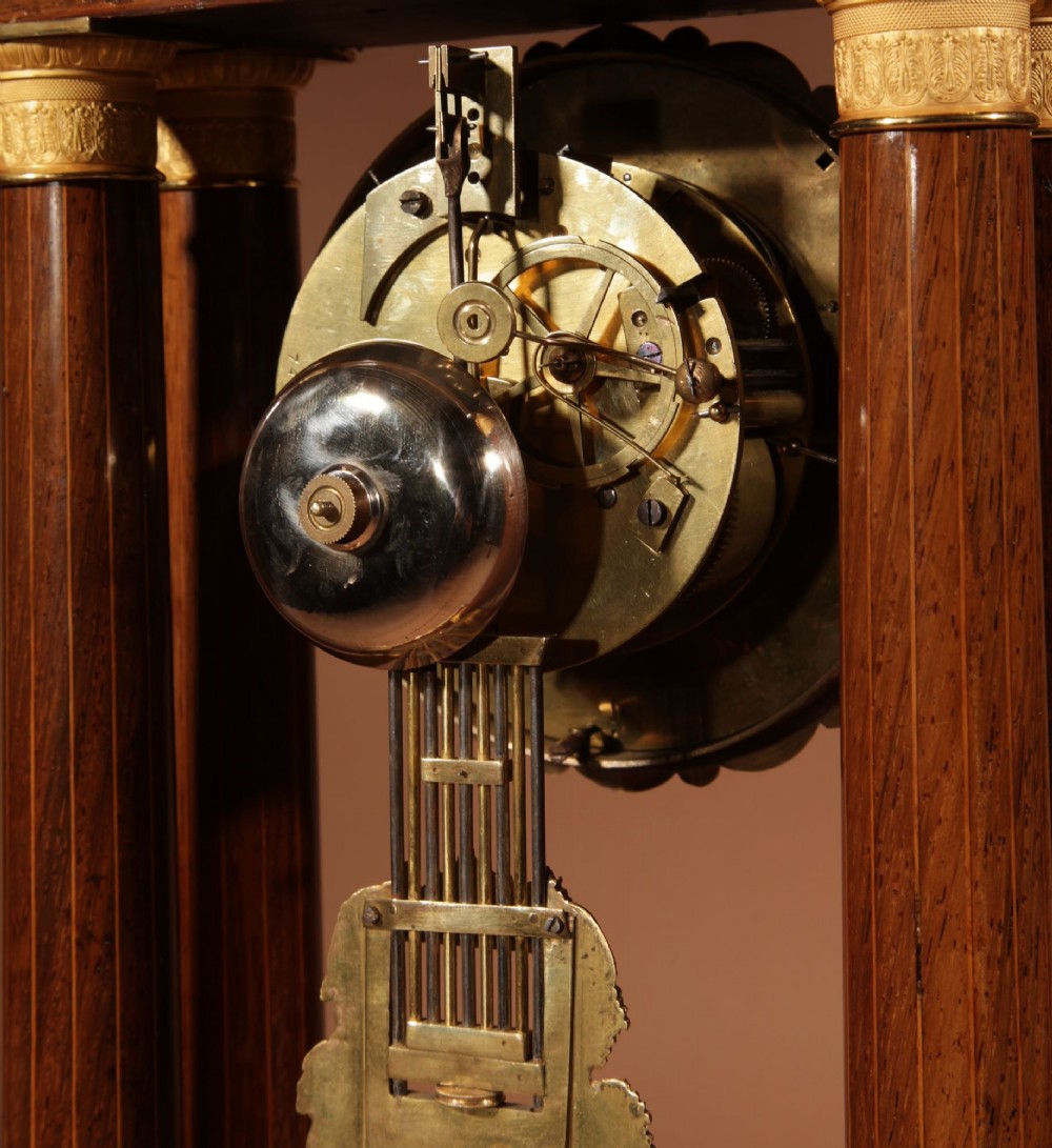 An antique rosewood portico clock with four fluted columns, gilt ormolu capitals, floral lemonwood marquetry inlays, and an exposed pendulum. View XI