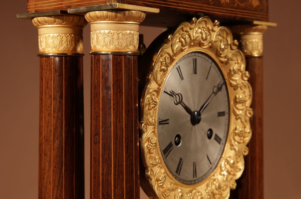 An antique rosewood portico clock with four fluted columns, gilt ormolu capitals, floral lemonwood marquetry inlays, and an exposed pendulum. View XII