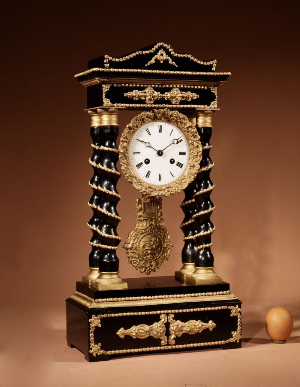 An ebonised wooden portico clock featuring four spiral columns with gilded mounts framing a central white enamel dial and decorative pendulum.