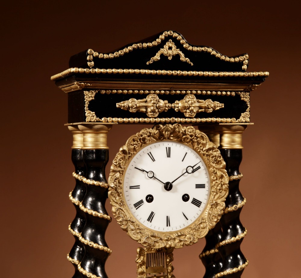 An ebonised wooden portico clock featuring four spiral columns with gilded mounts framing a central white enamel dial and decorative pendulum. View II