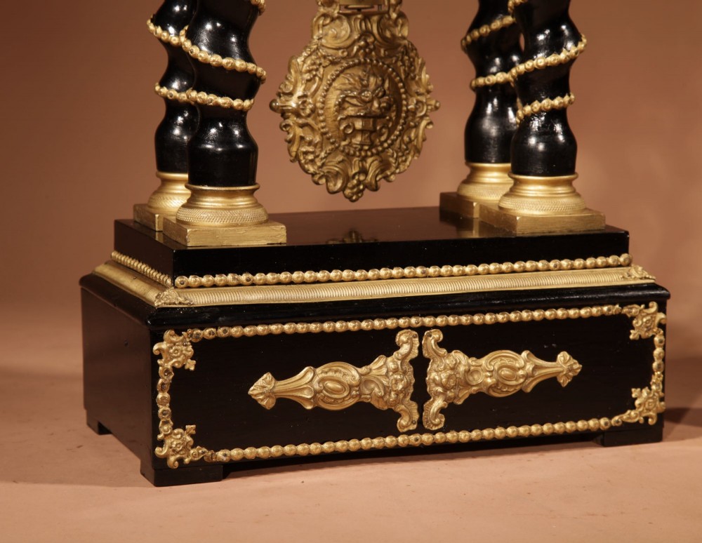 An ebonised wooden portico clock featuring four spiral columns with gilded mounts framing a central white enamel dial and decorative pendulum. View III