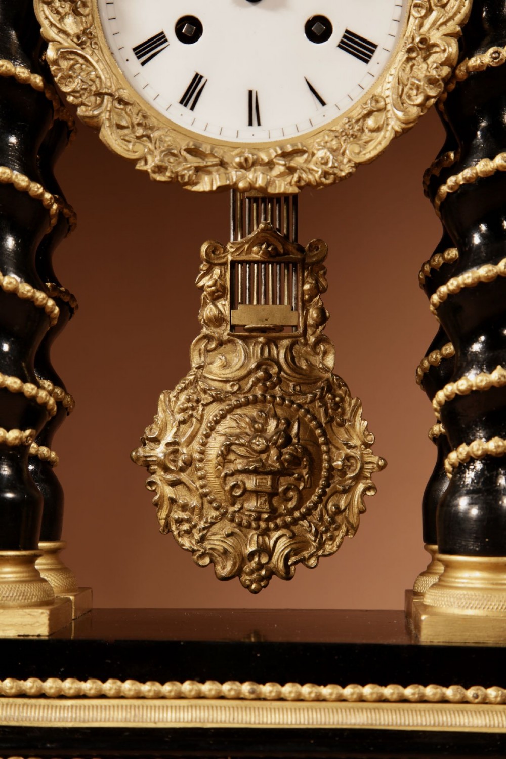 An ebonised wooden portico clock featuring four spiral columns with gilded mounts framing a central white enamel dial and decorative pendulum. View V
