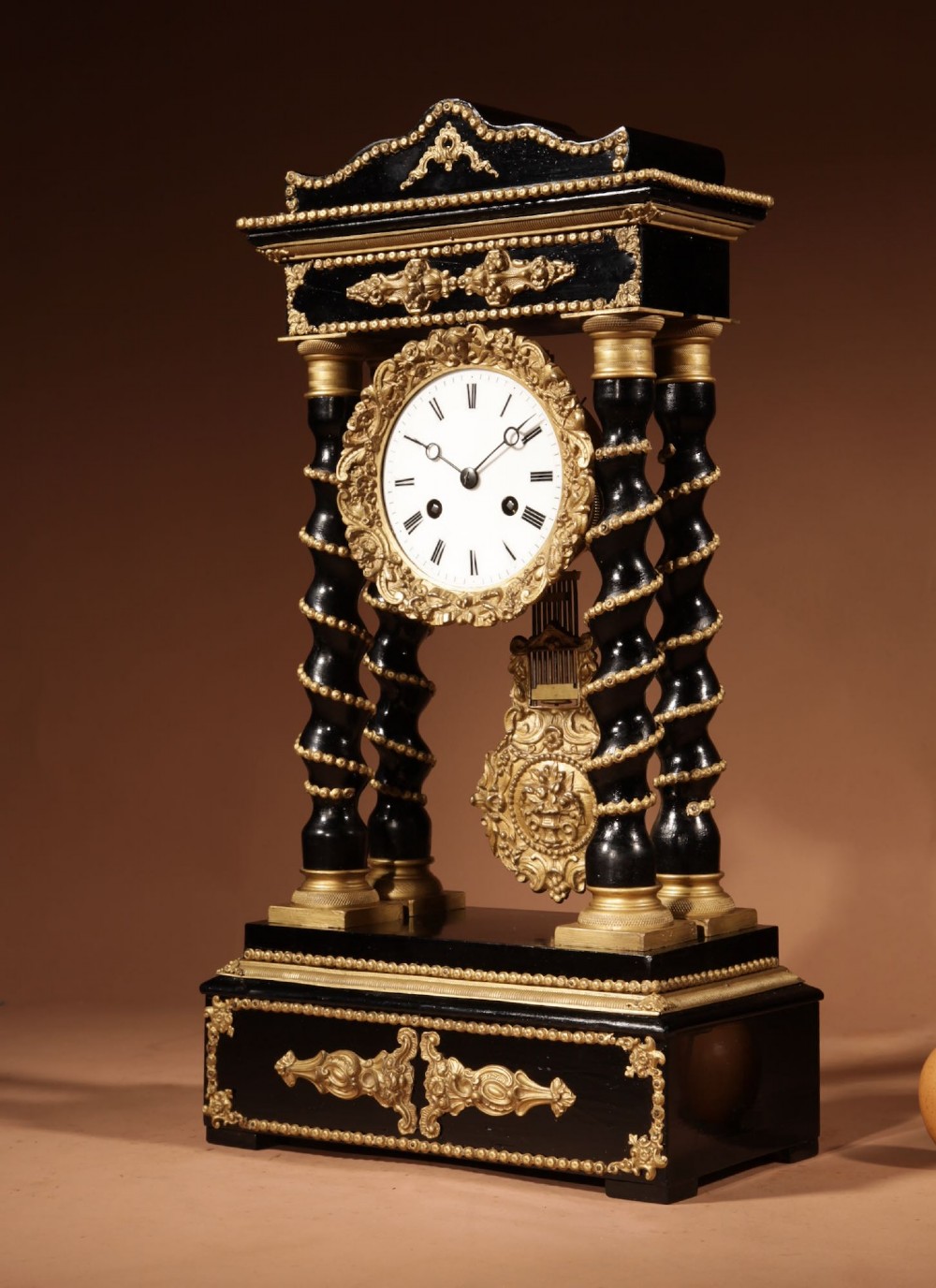 An ebonised wooden portico clock featuring four spiral columns with gilded mounts framing a central white enamel dial and decorative pendulum. View VI