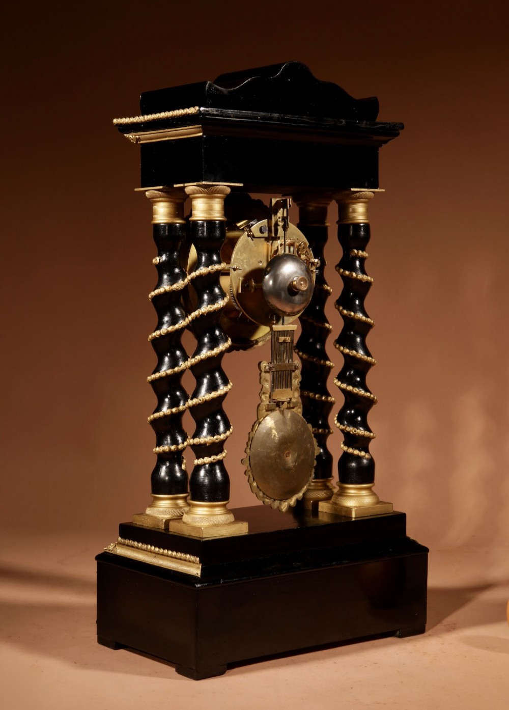 An ebonised wooden portico clock featuring four spiral columns with gilded mounts framing a central white enamel dial and decorative pendulum. View VII