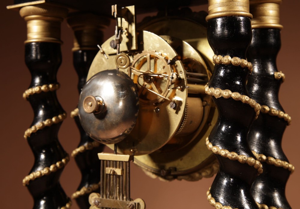 An ebonised wooden portico clock featuring four spiral columns with gilded mounts framing a central white enamel dial and decorative pendulum. View VIII