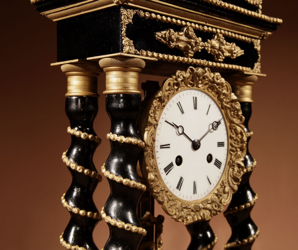 An ebonised wooden portico clock featuring four spiral columns with gilded mounts framing a central white enamel dial and decorative pendulum. View IX