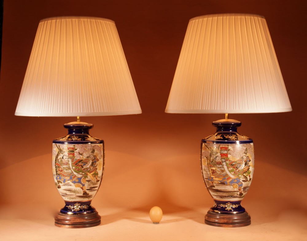 A pair of Japanese Satsuma ceramic table lamps featuring hand-painted figure scenes, cobalt blue accents, dark wooden bases, and cream pleated lampshades.