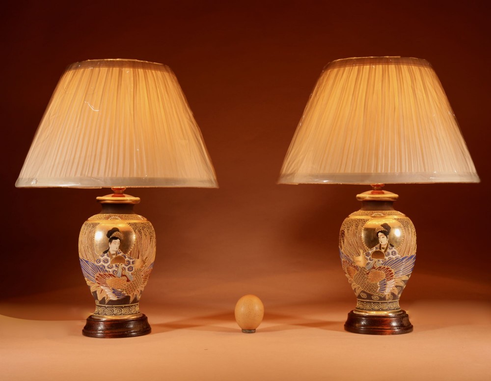 A pair of small Japanese Satsuma ceramic table lamps featuring figure paintings, gold gilding, dark wooden bases, and pleated cream shades.
