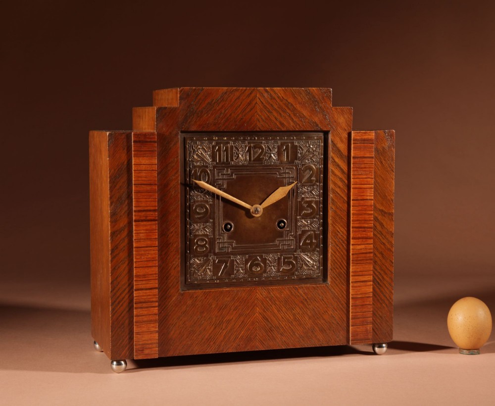 An Art Deco mantel clock crafted from oak and rosewood veneer featuring a stepped architectural top, a square brass dial with relief numerals, and small metal ball feet.