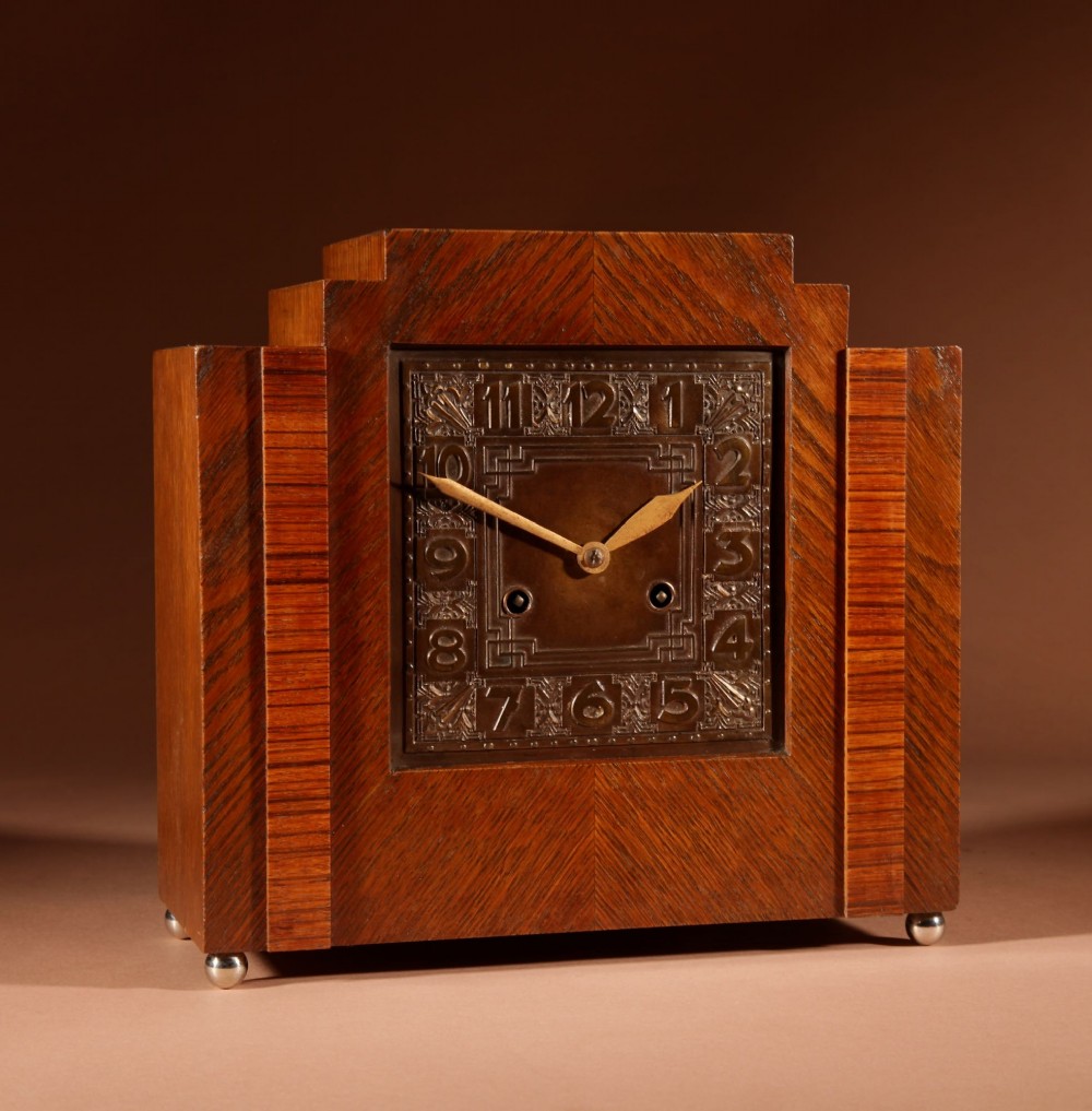 An Art Deco mantel clock crafted from oak and rosewood veneer featuring a stepped architectural top, a square brass dial with relief numerals, and small metal ball feet. View II