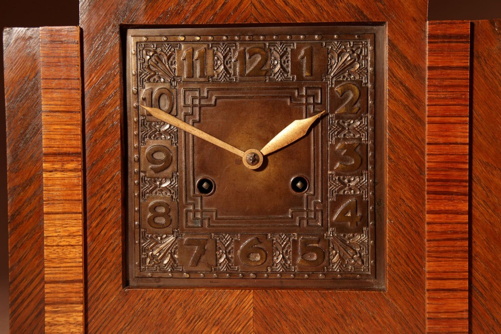 An Art Deco mantel clock crafted from oak and rosewood veneer featuring a stepped architectural top, a square brass dial with relief numerals, and small metal ball feet. View III