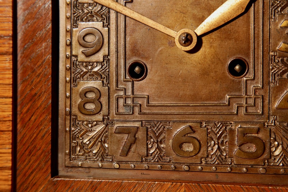 An Art Deco mantel clock crafted from oak and rosewood veneer featuring a stepped architectural top, a square brass dial with relief numerals, and small metal ball feet. View IV