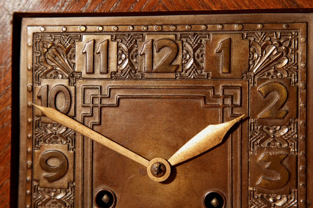 An Art Deco mantel clock crafted from oak and rosewood veneer featuring a stepped architectural top, a square brass dial with relief numerals, and small metal ball feet. View V
