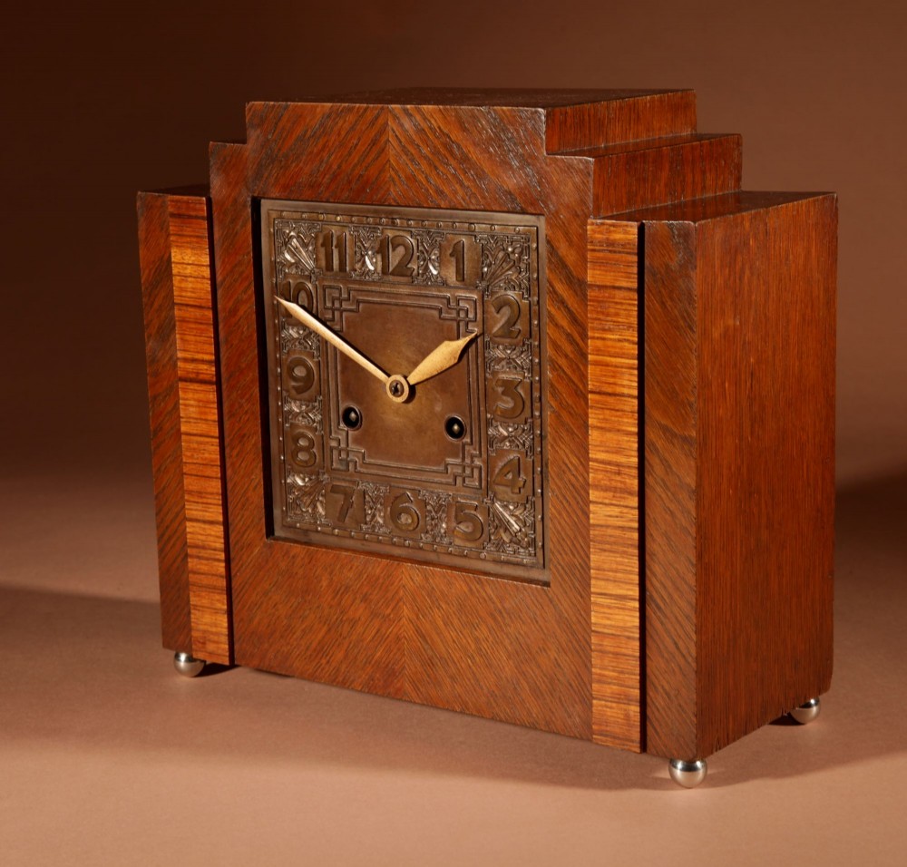 An Art Deco mantel clock crafted from oak and rosewood veneer featuring a stepped architectural top, a square brass dial with relief numerals, and small metal ball feet. View VI