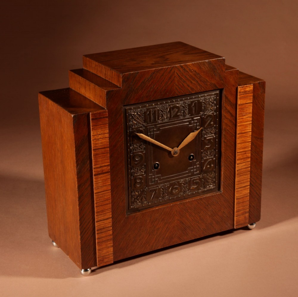 An Art Deco mantel clock crafted from oak and rosewood veneer featuring a stepped architectural top, a square brass dial with relief numerals, and small metal ball feet. View VII