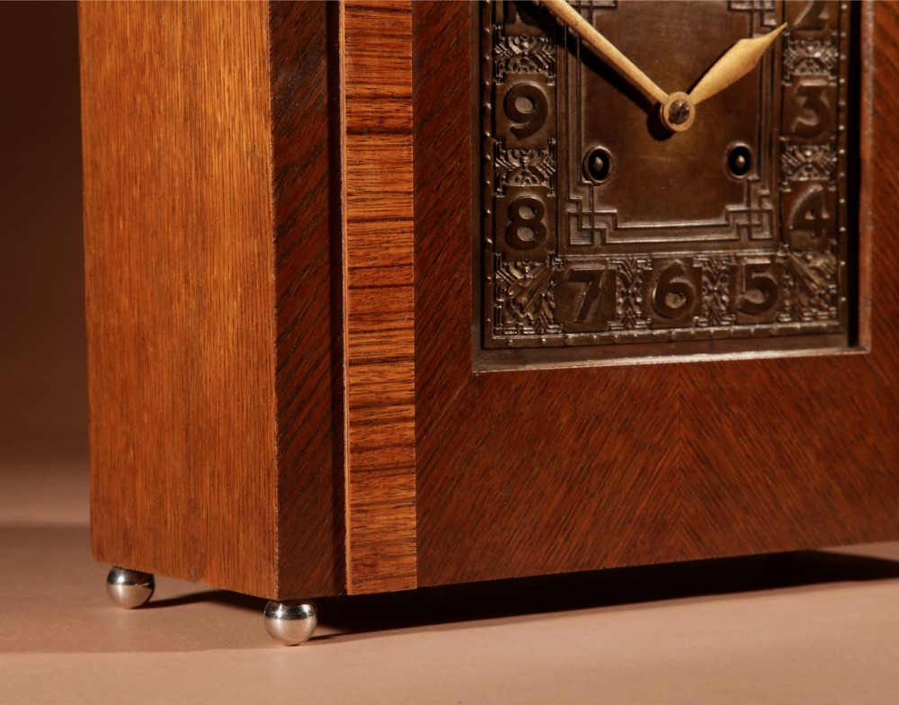 An Art Deco mantel clock crafted from oak and rosewood veneer featuring a stepped architectural top, a square brass dial with relief numerals, and small metal ball feet. View VIII