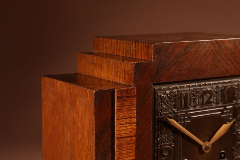 An Art Deco mantel clock crafted from oak and rosewood veneer featuring a stepped architectural top, a square brass dial with relief numerals, and small metal ball feet. View IX