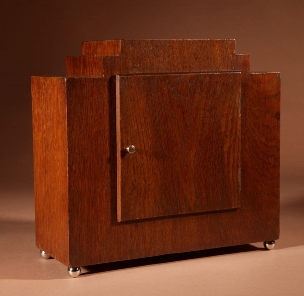An Art Deco mantel clock crafted from oak and rosewood veneer featuring a stepped architectural top, a square brass dial with relief numerals, and small metal ball feet. View X