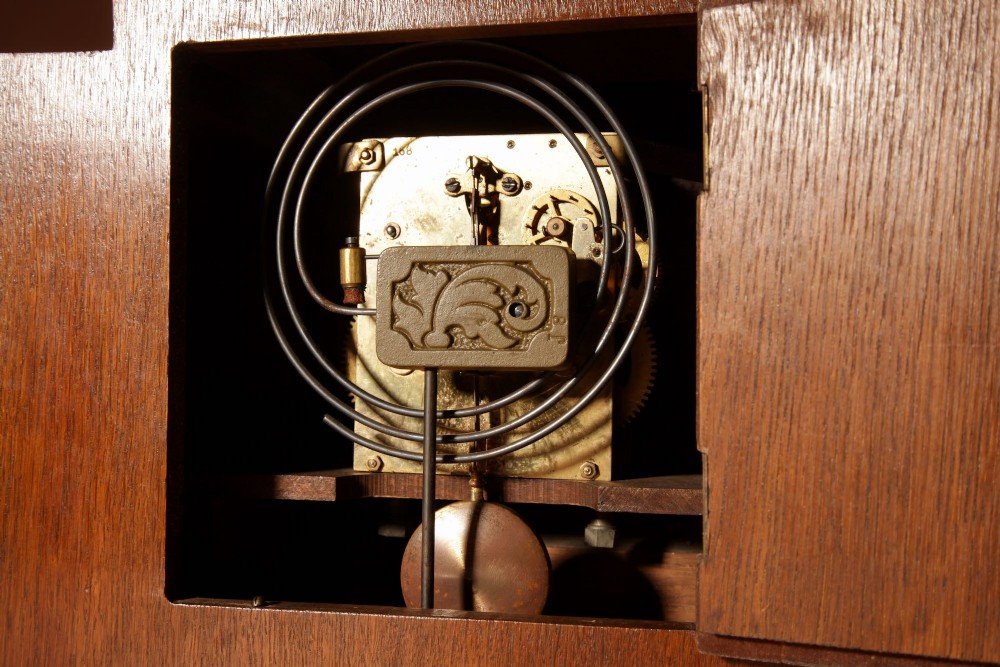 An Art Deco mantel clock crafted from oak and rosewood veneer featuring a stepped architectural top, a square brass dial with relief numerals, and small metal ball feet. View XI