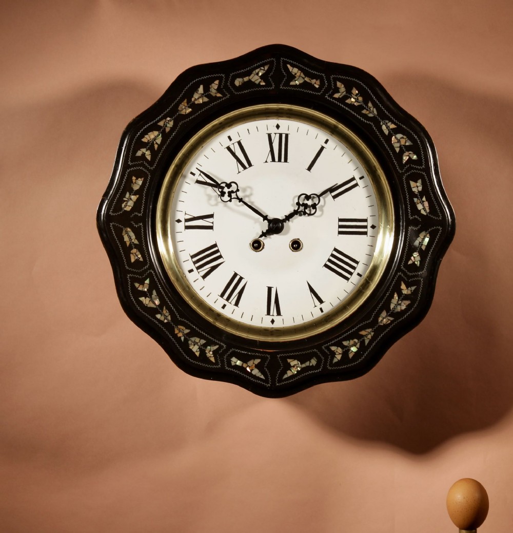 An ebonised French wall clock featuring a scalloped outer frame with floral mother-of-pearl inlay, a brass bezel, and a white dial with black Roman numerals.