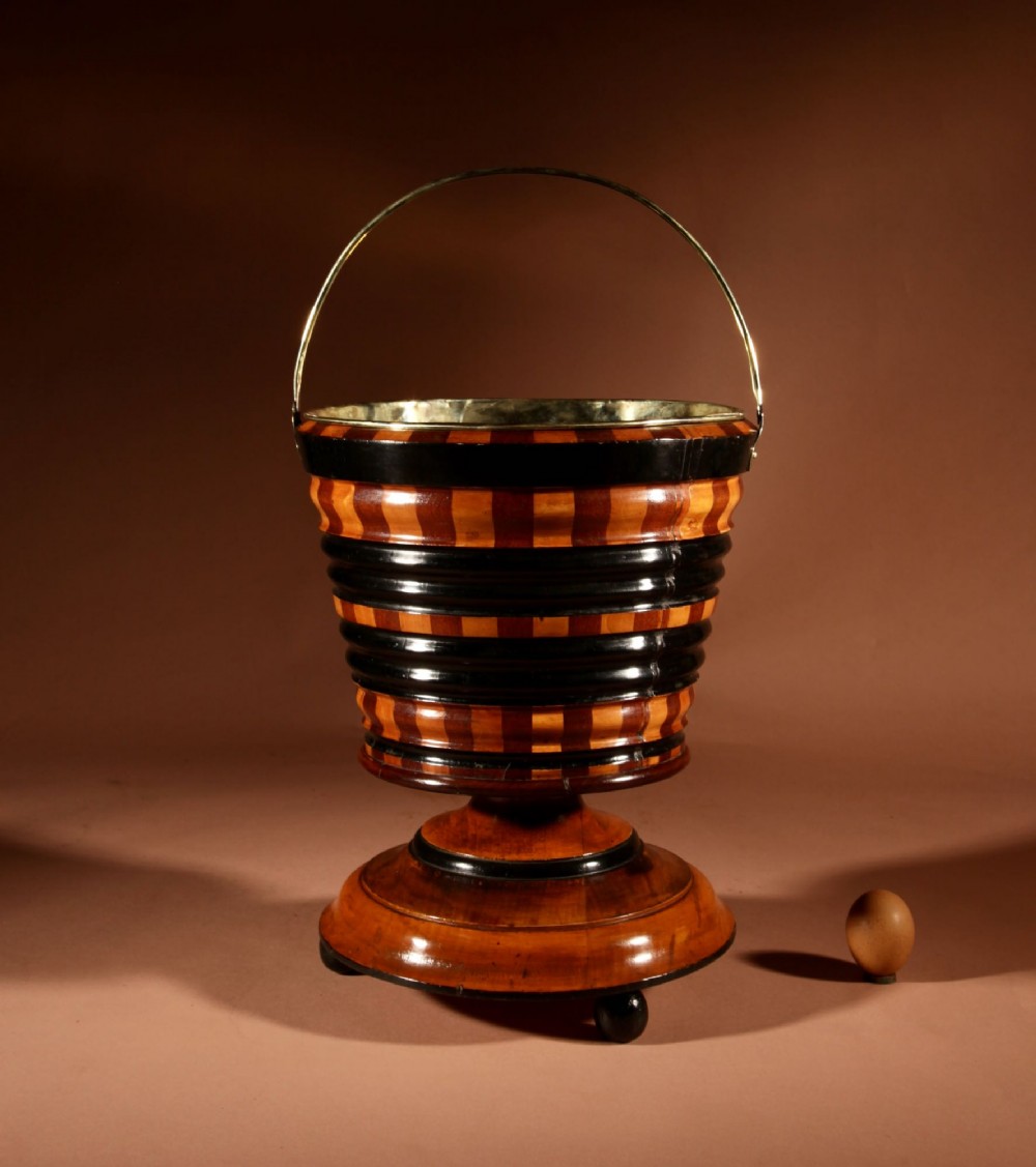 A Dutch coopered wooden peat bucket with alternating brown and ebonised staves, a brass swing handle, and a removable metal liner on a footed pedestal base.