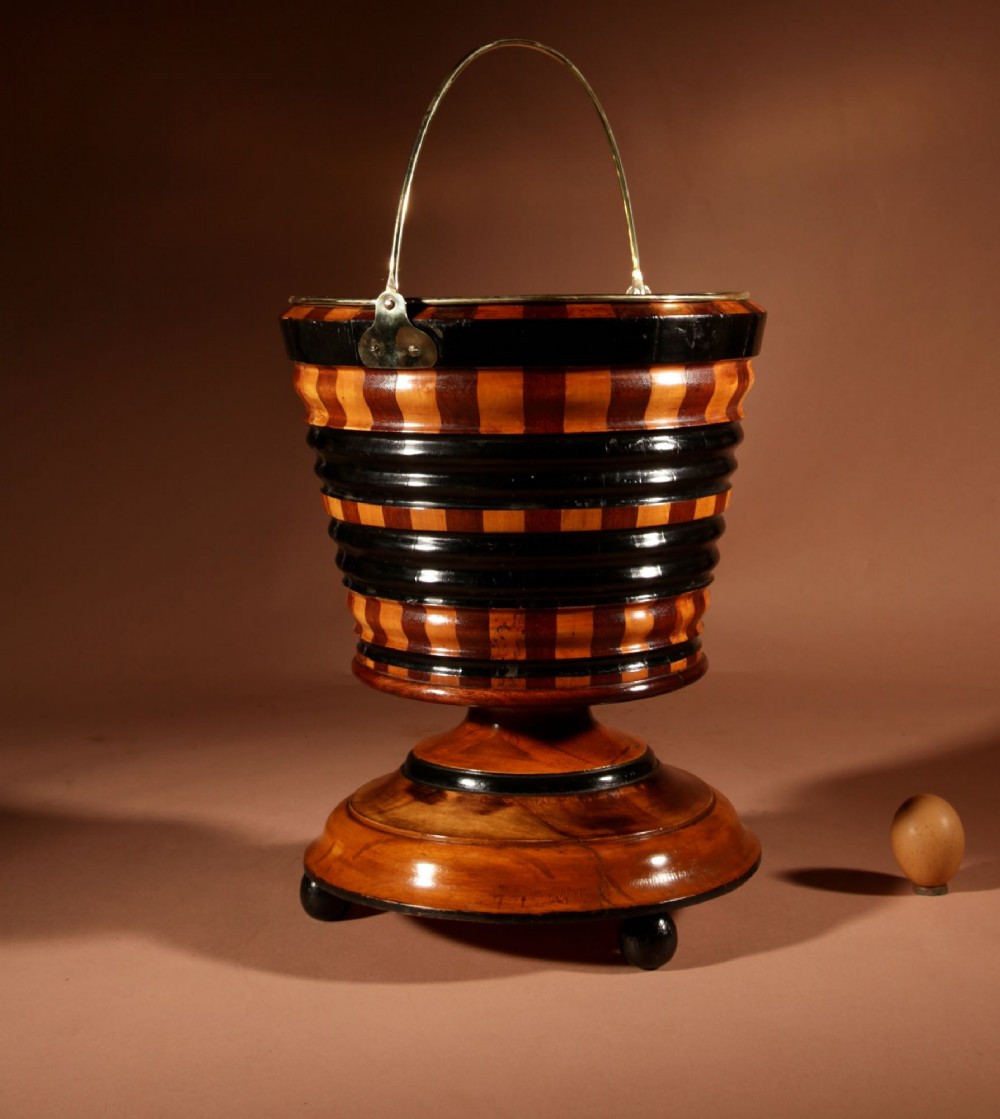 A Dutch coopered wooden peat bucket with alternating brown and ebonised staves, a brass swing handle, and a removable metal liner on a footed pedestal base. View III