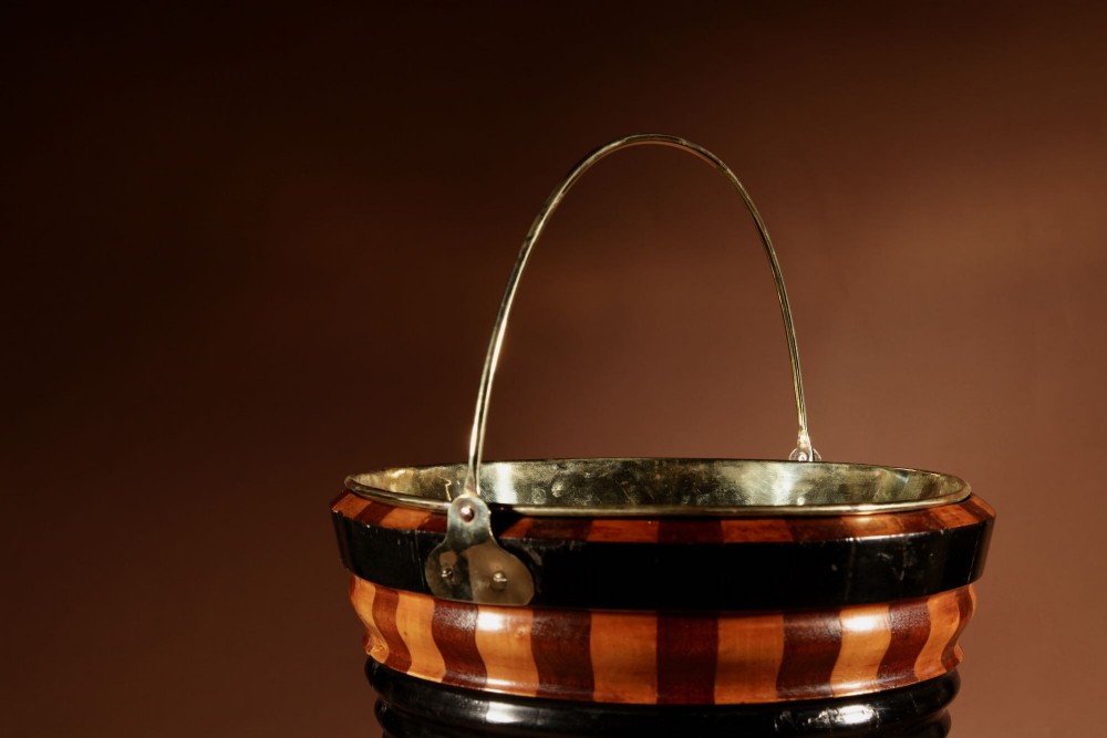 A Dutch coopered wooden peat bucket with alternating brown and ebonised staves, a brass swing handle, and a removable metal liner on a footed pedestal base. View IV