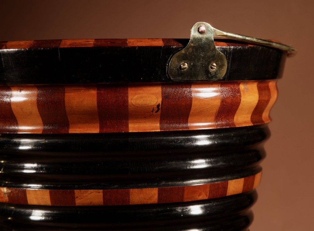 A Dutch coopered wooden peat bucket with alternating brown and ebonised staves, a brass swing handle, and a removable metal liner on a footed pedestal base. View IX