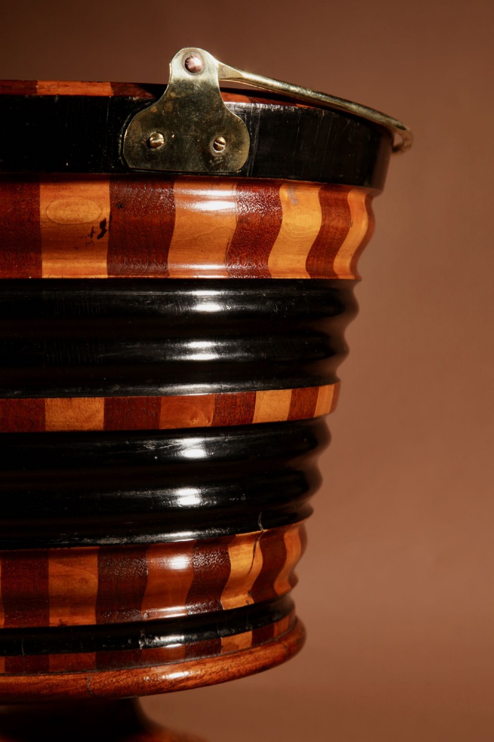 A Dutch coopered wooden peat bucket with alternating brown and ebonised staves, a brass swing handle, and a removable metal liner on a footed pedestal base. View X