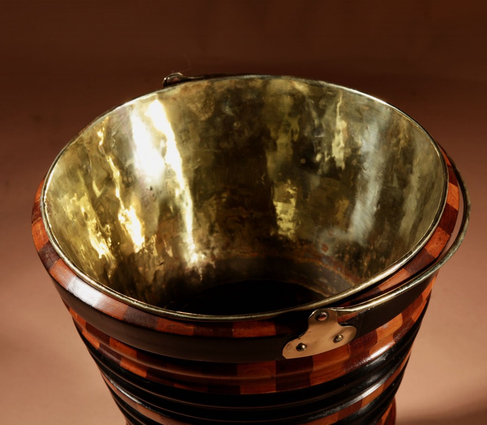 A Dutch coopered wooden peat bucket with alternating brown and ebonised staves, a brass swing handle, and a removable metal liner on a footed pedestal base. View XI