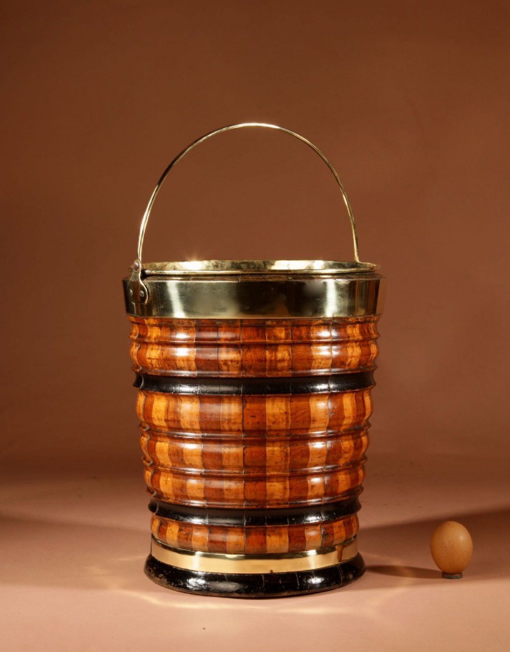 An antique coopered wooden peat bucket featuring alternating light and dark ribbed wood staves, ebonised bands, a brass rim, and a brass swing handle.