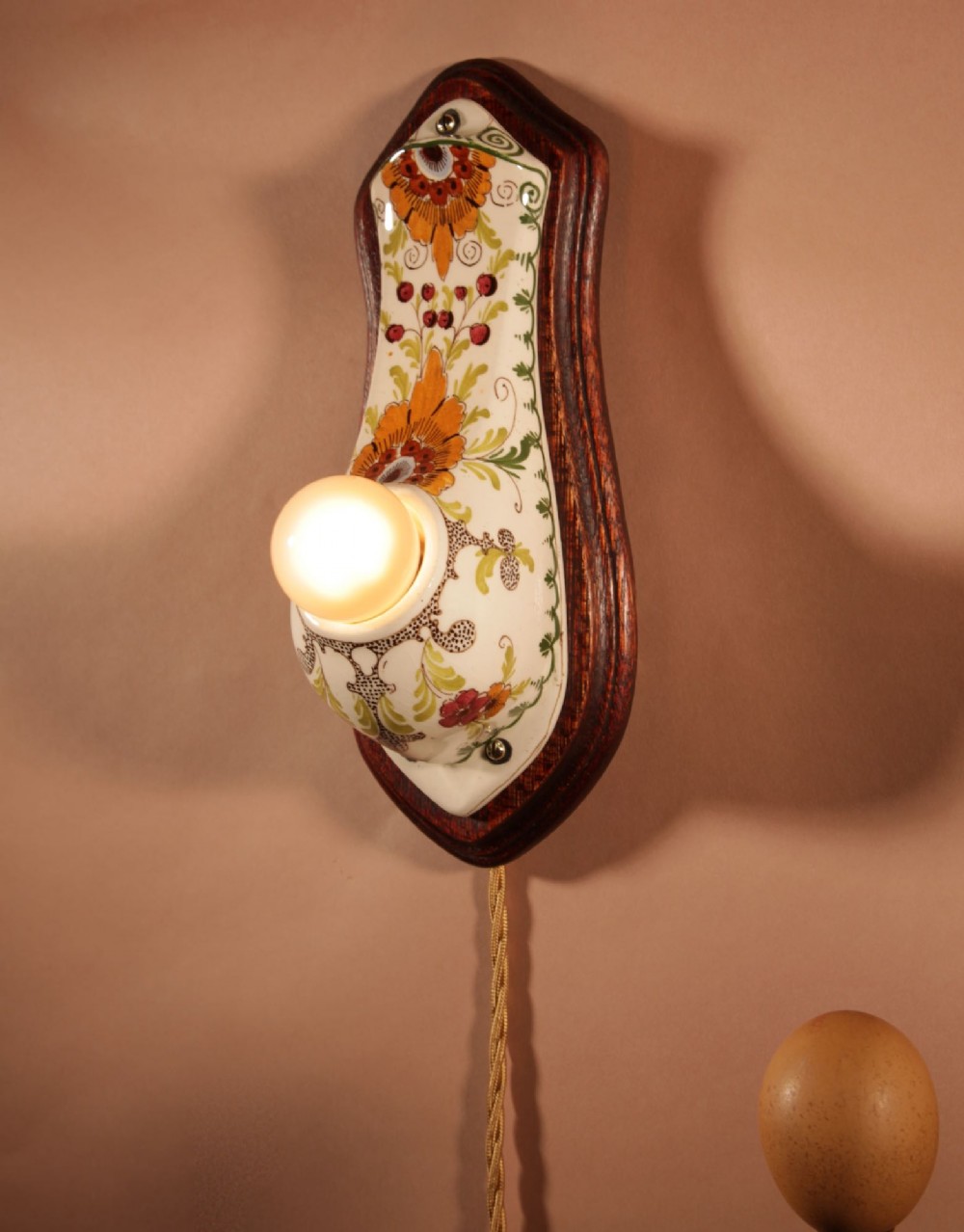 A Dutch Art Nouveau hand-painted ceramic wall sconce with floral patterns, mounted on a dark wooden backplate with an illuminated bulb. View II