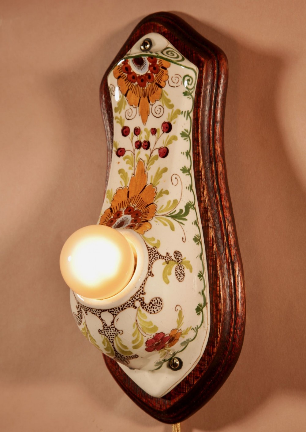 A Dutch Art Nouveau hand-painted ceramic wall sconce with floral patterns, mounted on a dark wooden backplate with an illuminated bulb.