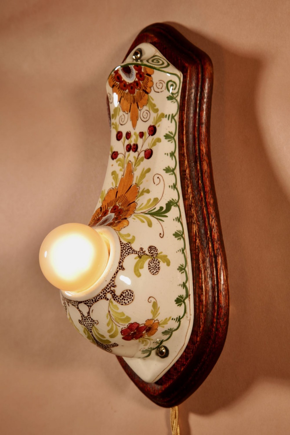 A Dutch Art Nouveau hand-painted ceramic wall sconce with floral patterns, mounted on a dark wooden backplate with an illuminated bulb. View III