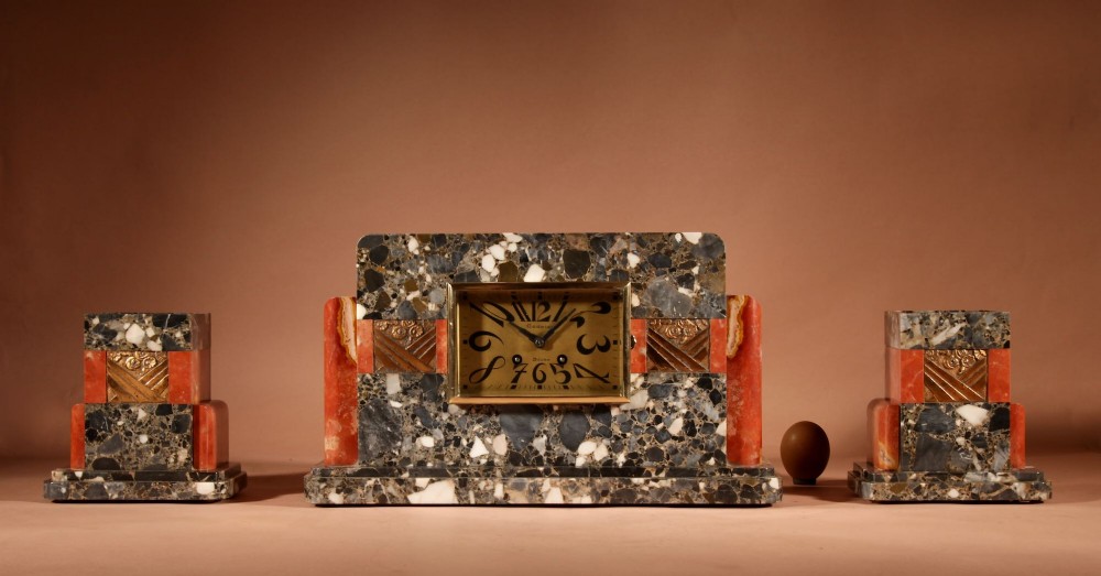 An Art Deco clock garniture set constructed from dark speckled stone and coral marble featuring a central rectangular clock with gilt numerals and two matching side pieces.