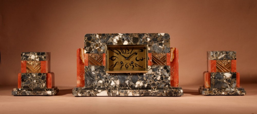 An Art Deco clock garniture set constructed from dark speckled stone and coral marble featuring a central rectangular clock with gilt numerals and two matching side pieces. View II