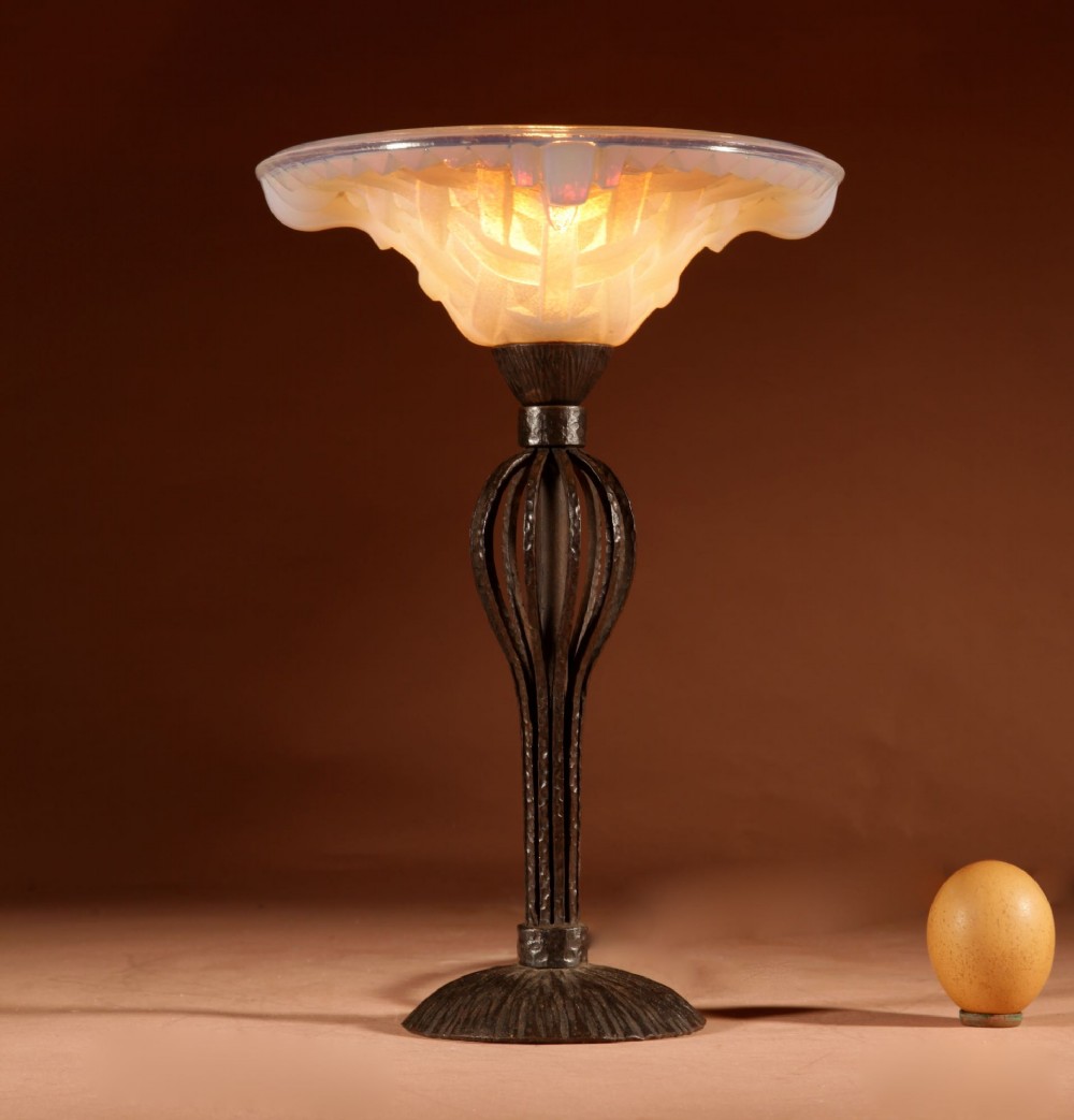 An illuminated French Art Deco table lamp with a flared opalescent glass shade set atop a dark, hammered wrought iron stem and circular base.