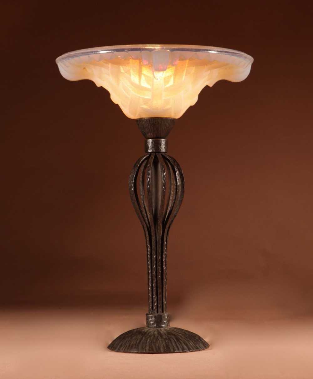 An illuminated French Art Deco table lamp with a flared opalescent glass shade set atop a dark, hammered wrought iron stem and circular base. View II